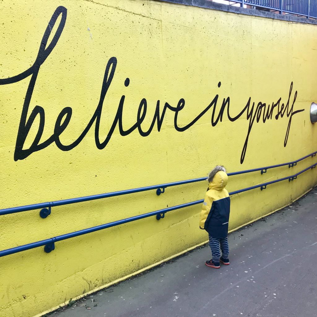 A child looks up at a yellow wall. On the wall we see the words "believe in yourself" in large black cursive writing. 