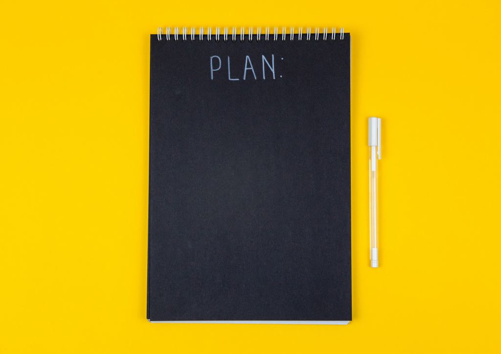A spiral-bound planner, with a pen beside it, rests on a yellow background 