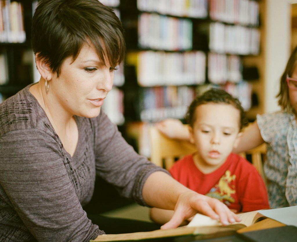A woman sits at a desk and reads to two small children. There are several bookshelves in the background. 
