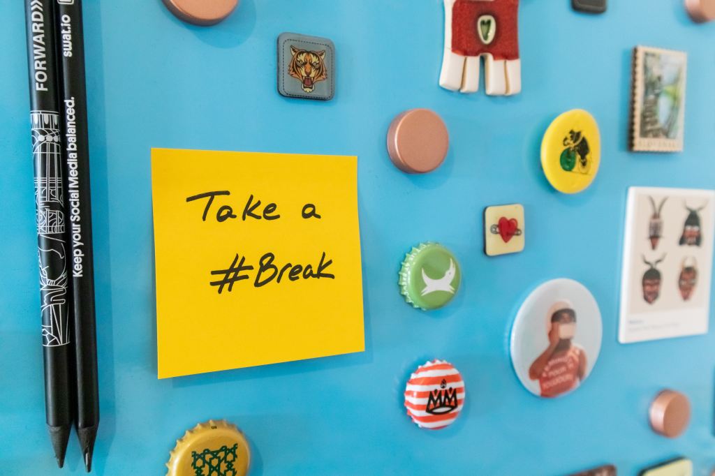 A blue bulletin board with various items stuck on it, including a bright yellow post-it note with the words "Take a #Break." 