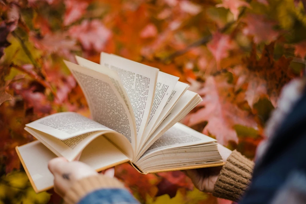 A book is held open with colourful (out-of-focus) leaves in the background.